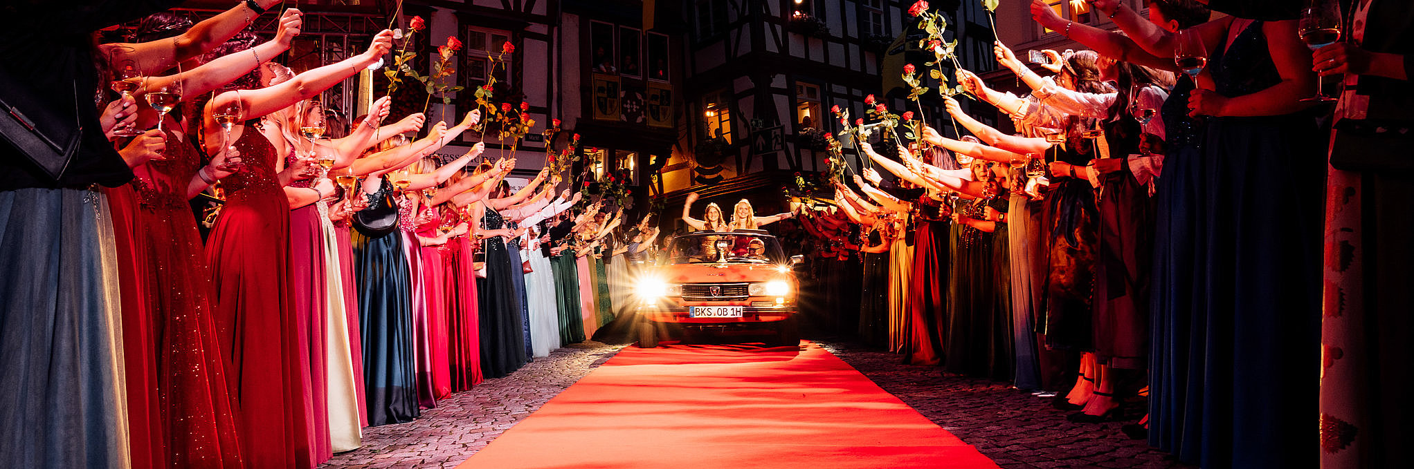 Empfang der Mosella am Weinfest der Mittelmosel in Bernkastel-Kues. Weinhoheiten aus den Moselorten stehen links und rechts entlang eines roten Teppichs Spalier und halten Rosen in der Hand. In der Mitte kommen auf einem roten Oldtimer die Mosella und Weinprinzessin von Bernkastel-Kues eingefahren.