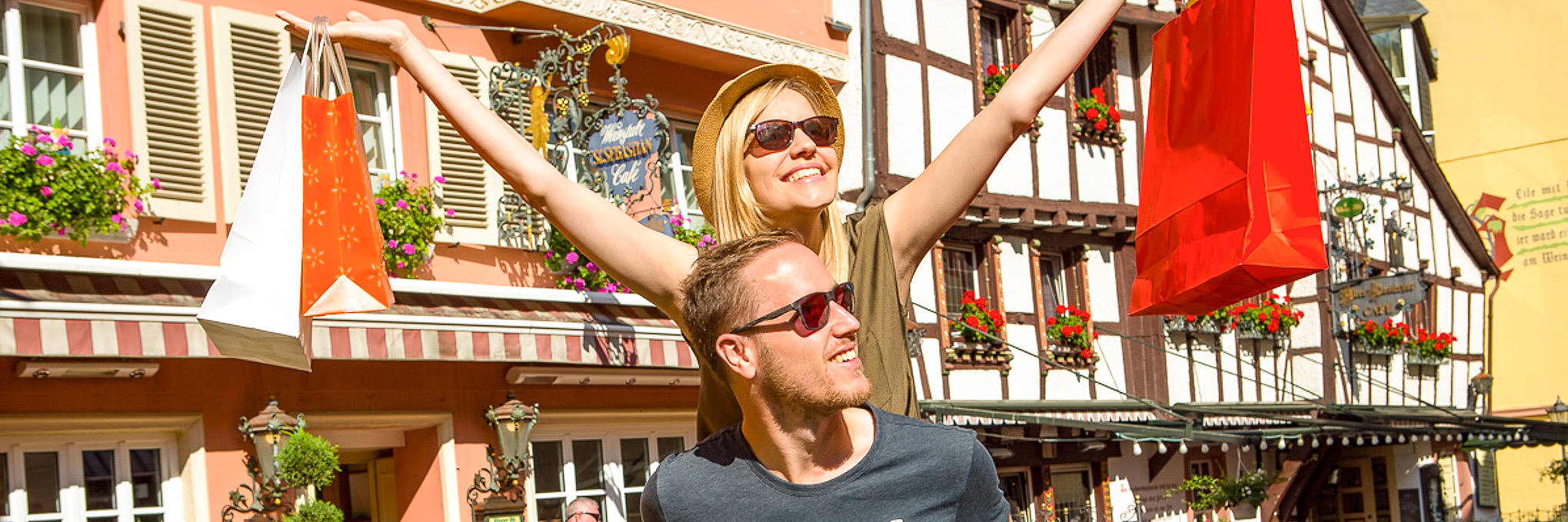 Zwei Personen beim Shopping in der Altstadt von Bernkastel-Kues Das Foto zeigt zwei Personen beim Shopping in Bernkastel-Kues. Die eine Person trägt die andere Person auf dem Rücken, während die die Shoppingtaschen in die Höhe hält. Im Hintergrund ist die historische Bebauung der Stadt zu sehen.