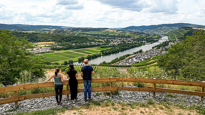 Das Foto zeigt drei Personen, die mit dem Rücken zur Kamera stehen. Die Personen stehen an einem Aussichtspunkt und schauen ins Moseltal. Zu sehen sind die Weinberge, Ortschaften und die Mosel.