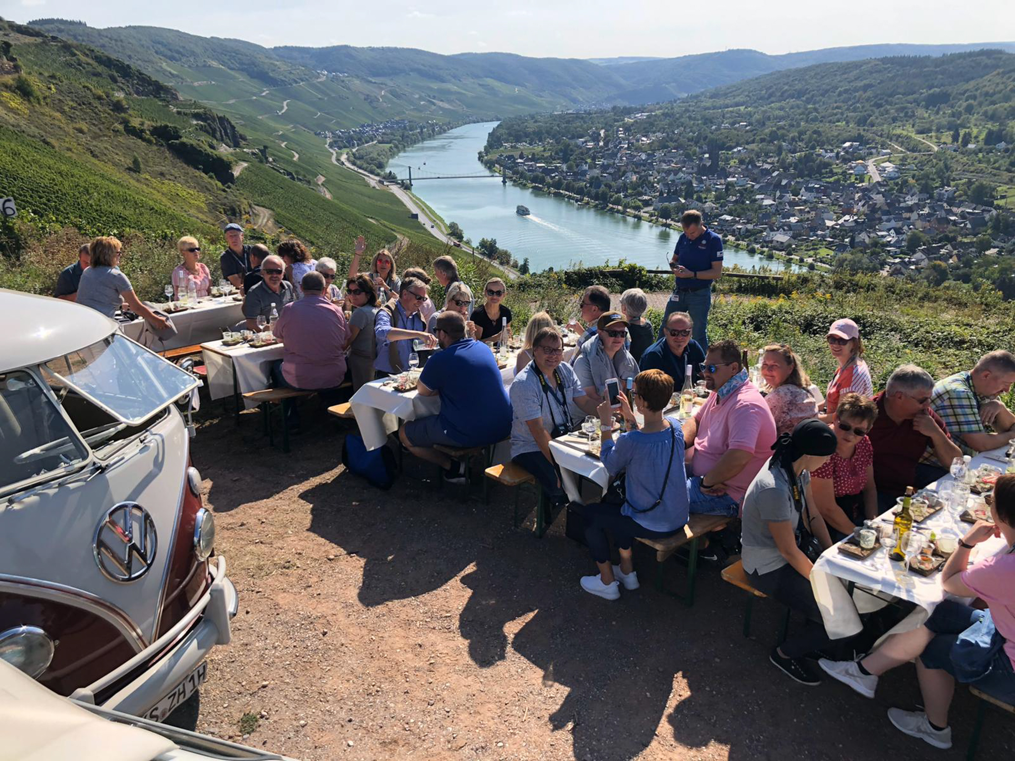 Nostalgische Bulli-Tour an der Mosel mit Picknick im Weinberg Das Bild zeigt ein Oldtimer VW Bulli in den Weinbergen. Davor sitzen viele Leute auf Bänken und sind am Essen und am Trinken. Im Hintergrund fließt die Mosel.