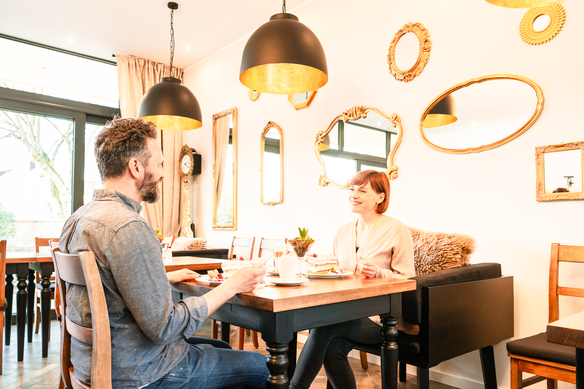 Zwei Personen beim Cafébesuch in Bernkastel-Kues Das Foto zeigt zwei Personen, die ein Café besuchen. Sie genießen ein Stück Kuchen und trinken einen Kaffee. An der Wand des Cafés hängen viele Spiegel mit goldenen Rahmen. Die Spiegel haben unterschiedliche Formen.